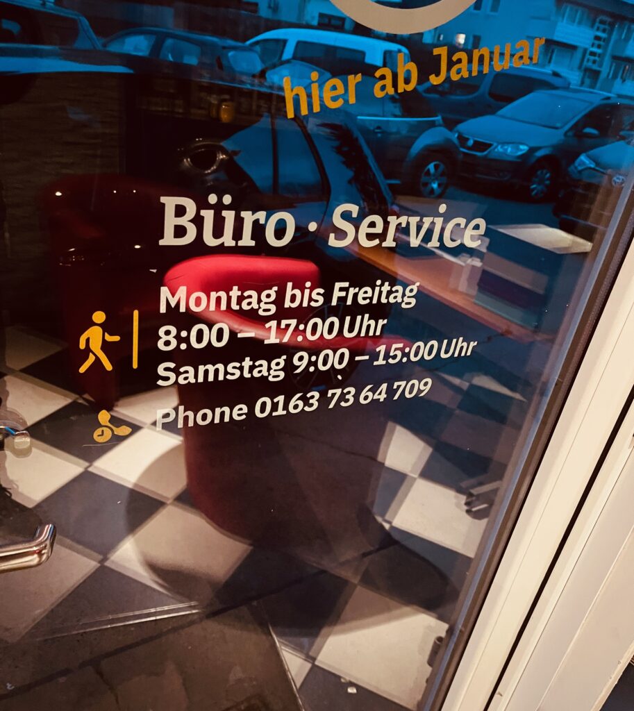 Glastür des HOCO Automarkts in der Marienstraße mit Büro-Service-Öffnungszeiten, Telefonnummer und Blick auf geparkte Gebrauchtwagen draußen